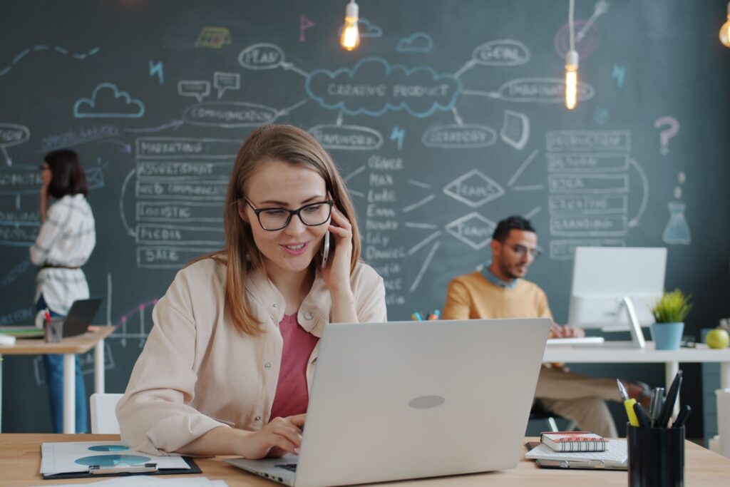 Business lady is speaking on mobile phone and taking notes in modern open space office while people are working in background. Communication and smartphone concept.